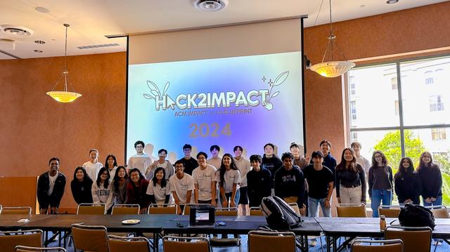 Over 20 people stand in front of a projected screen that says "Hack2Impact 2024"
